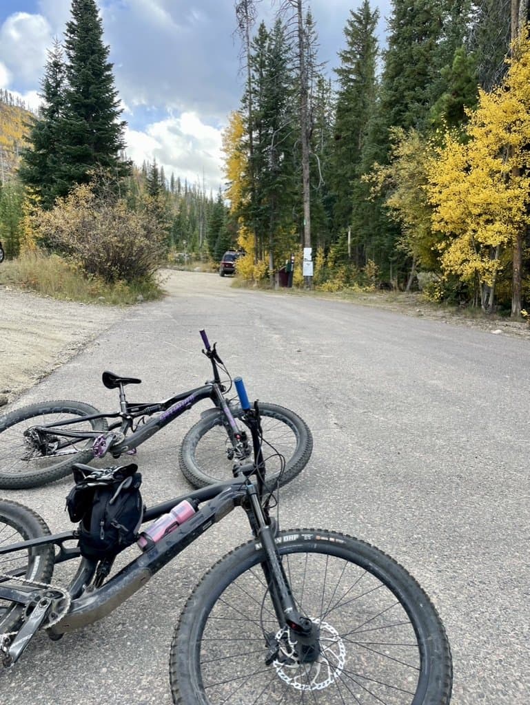Two mountain bikes resting on a gravel road at a trailhead, with yellow aspens and evergreens behind them