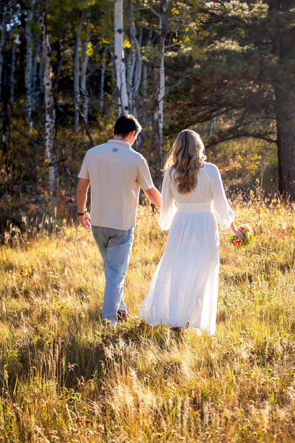 A couple walking hand in hand through a golden meadow toward aspen trees, the bride in a long white dress holding wildflowers